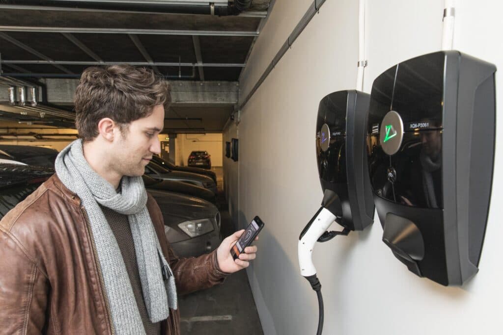 en man med brun skinnjacka och grå halsduk står med sin telefon framför två Zaptec Pro som är installerade i ett parkeringsgarage, en laddkabel är inkopplad i den ena laddaren och i bakgrunden syns bilar som står parkerade.