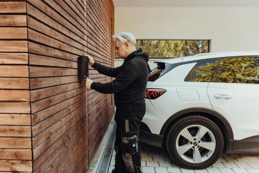 En man med silverfärgat hår installerar en Zaptec Pro laddbox på en trävägg bredvid en vit SUV på en uppfart till ett bostadshus.