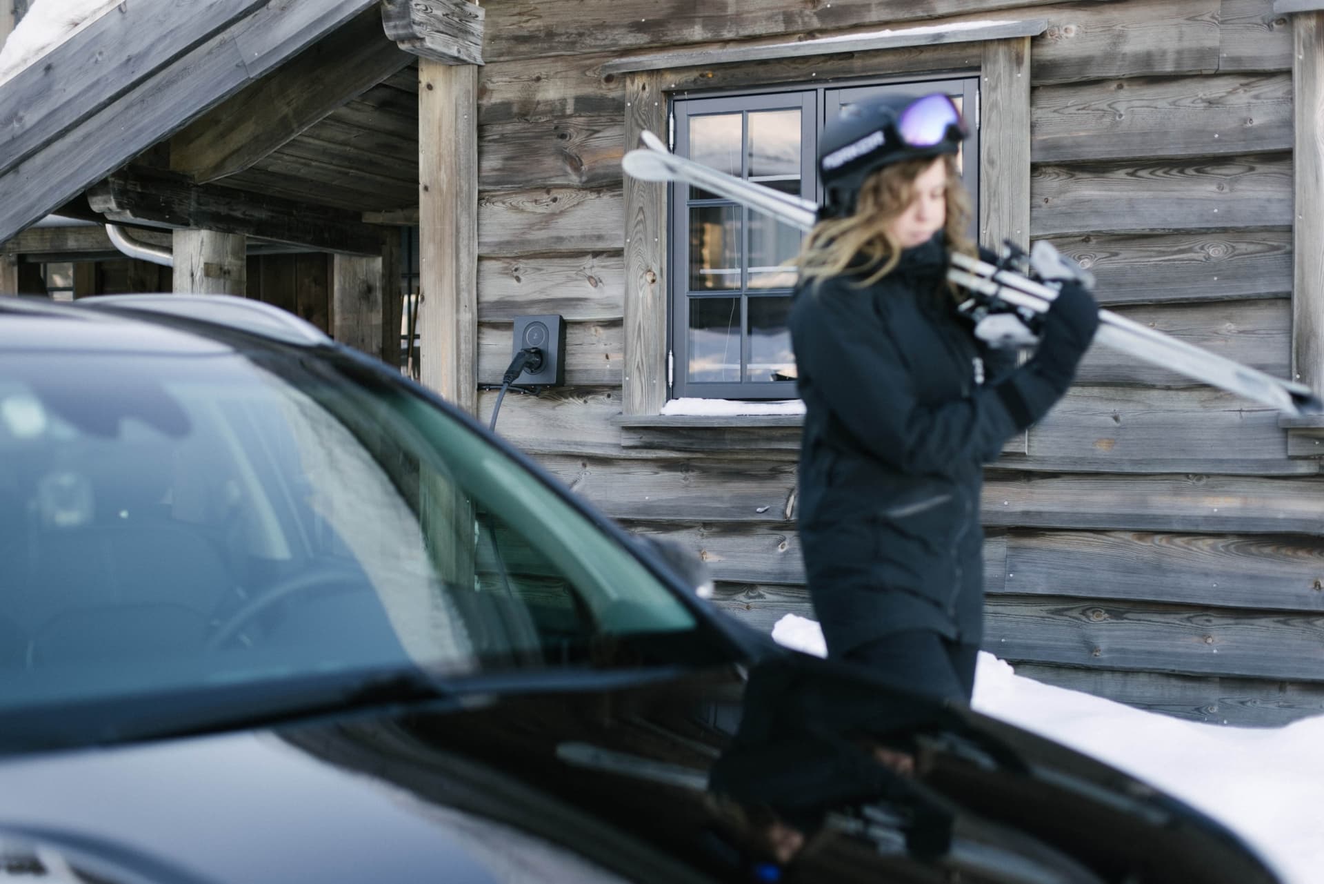 Kvinna som bär på skidor vid en timmerstuga. Framför henne står en svart bil som laddar via en svart Zapte Go laddare som sitter på väggen på det timmrade huset.