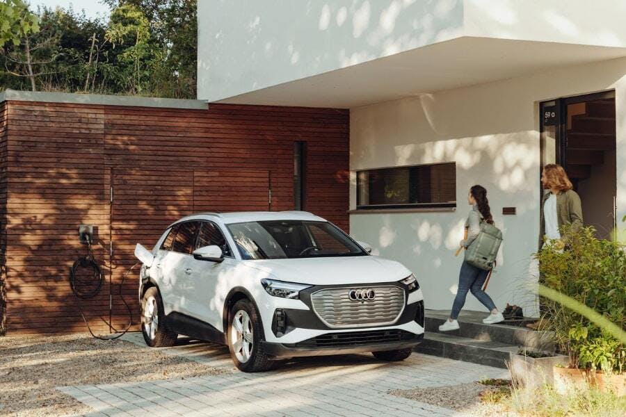 En vit Audi elbil står parkerad framför ett modernt hus med träpanel. Bilen är ansluten till en Zaptec laddstation på väggen. En kvinna och en man går mot bilen.