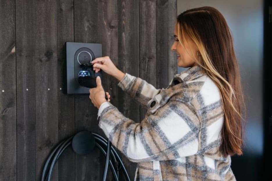 En glad kvinna iklädd en rutig jacka som stoppar i en laddkabel i en mörk grå Zaptec Go laddbox.