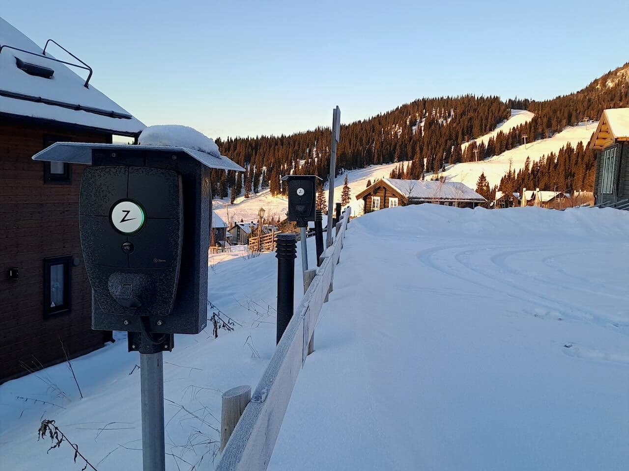 En snöig parkering med 4 Zaptec Pro omges av ett vinterlandskap med bruna stugor och ett berg med skidbackar lyses upp av solen