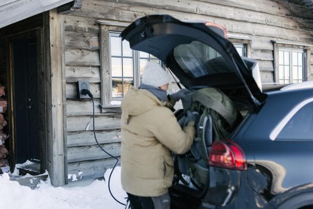 En person iklädd en varm jacka och mössa lastar ur utrustning från bagageutrymmet på en svart bil som står parkerad i snön. Bilen laddar med en Rock Grey Zaptec Go laddare monterad på den timrade väggen av en rustik stuga.