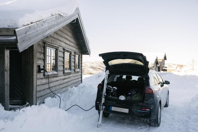 En mörkblå bil står parkerad utanför en trästuga i ett snötäckt landskap. Bilen laddas med en Zaptec Go-laddare monterad på stugans vägg. Bilens bagagelucka är öppen och ett par skidor står lutade mot bilen och annan utrustning är packad i bilen.