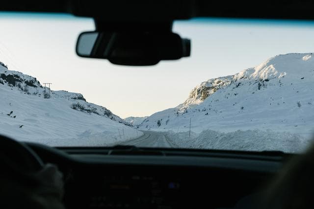 Utsikten från insidan av en bil som kör genom ett snötäckt bergspass. Vägen omges av höga snövallar och det sena solljuset lyser upp de karga topparna i fjärran.