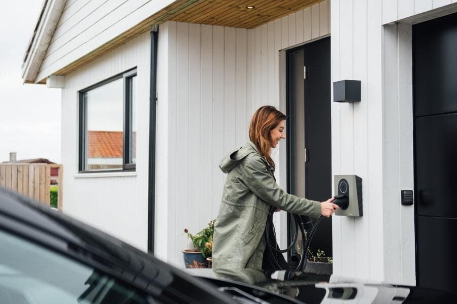 En kvinna laddar sin elbil hemma med en laddbox monterad på väggen vid entrén till ett hus. Hon bär en grön jacka och ser glad ut. Huset har vita träpaneler och ett modernt utseende.