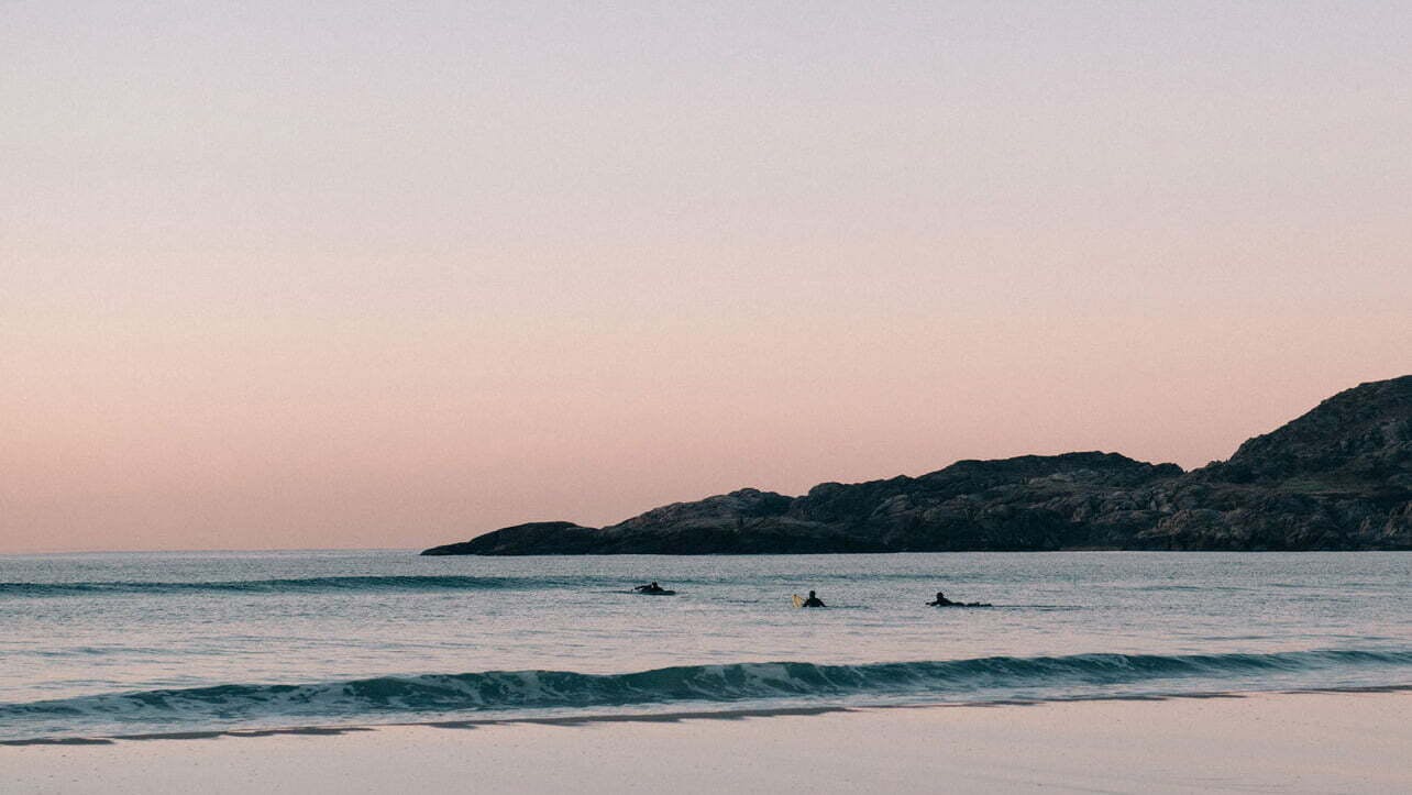 en strand med vågor som slår in, i vattnet är ste personer som simmar med surfbrädor, himlen går i rosa toner och steniga berg växer upp ut vattnet.