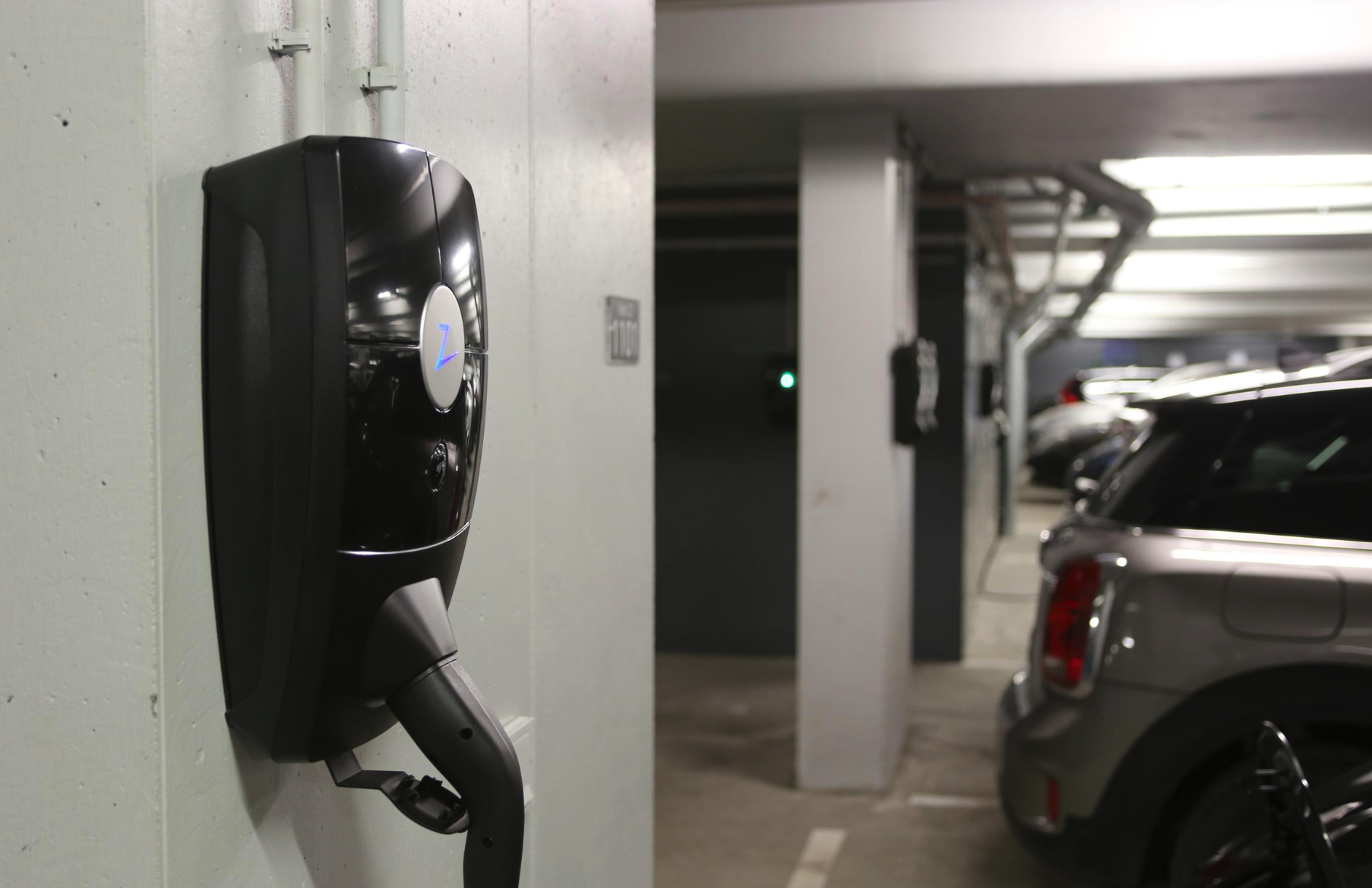 En elbilsladdare monterad på en vägg i ett underjordiskt parkeringsgarage. En silverfärgad bil är parkerad bredvid laddaren, och det finns fler bilar i bakgrunden