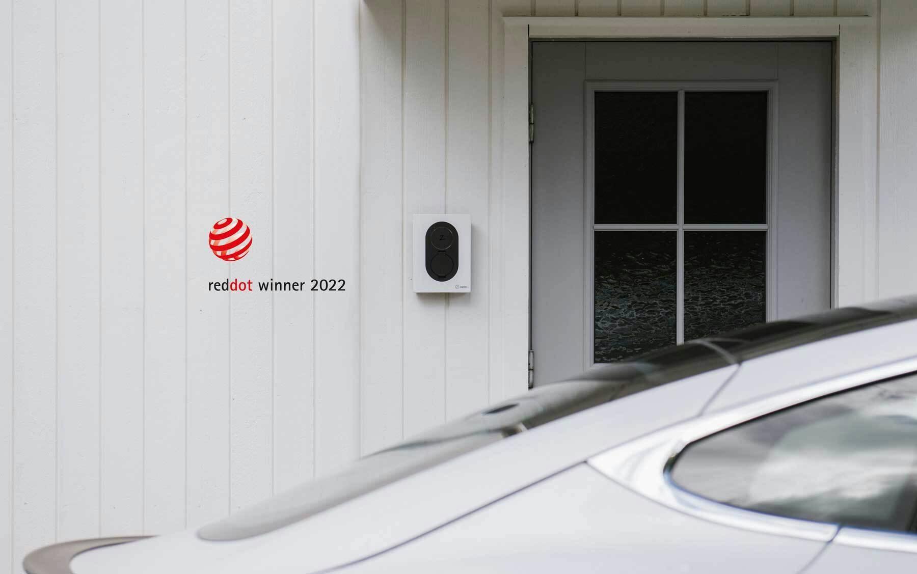 En vit Zaptec Go laddare sitter monterad på en vit träfasad bredvid en ljust grå dörr med mörka fönster, i förgrunden ser man delar av taket på en vit bil. I ena hörnet är loggan för reddot