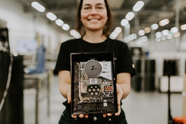 En glad kvinna med svart t-shirt och glasögon står i en ljus fabrik och håller fram en Zaptec Go laddare utan front cover.