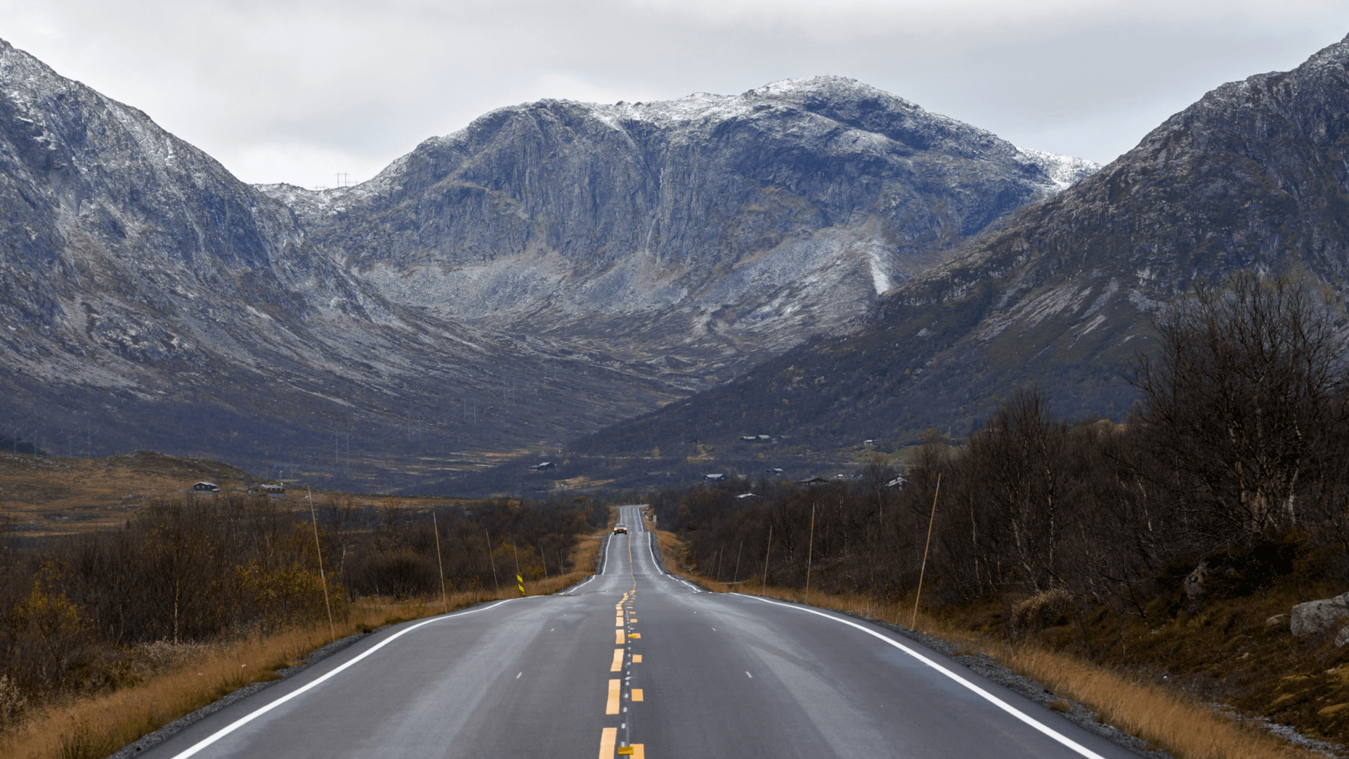En landsväg som går igenom ett mörkt landskap och omringas av grå berg med snö på toppen