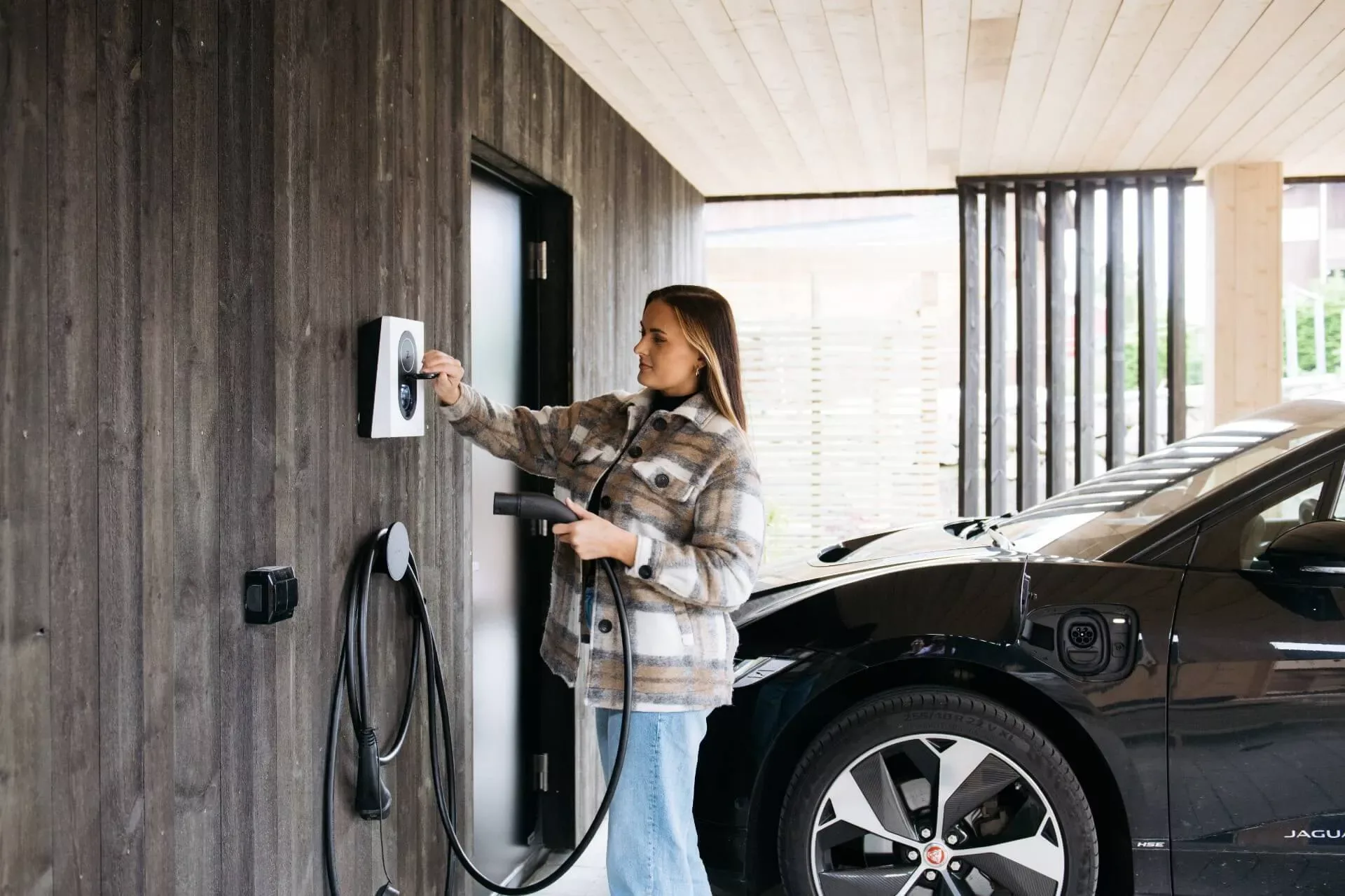 En kvinne i mønstret jakke kobler til en ladekabel i en Cloud White Zaptec Go-lader montert på en mørk trevegg, ved siden av en svart elbil.