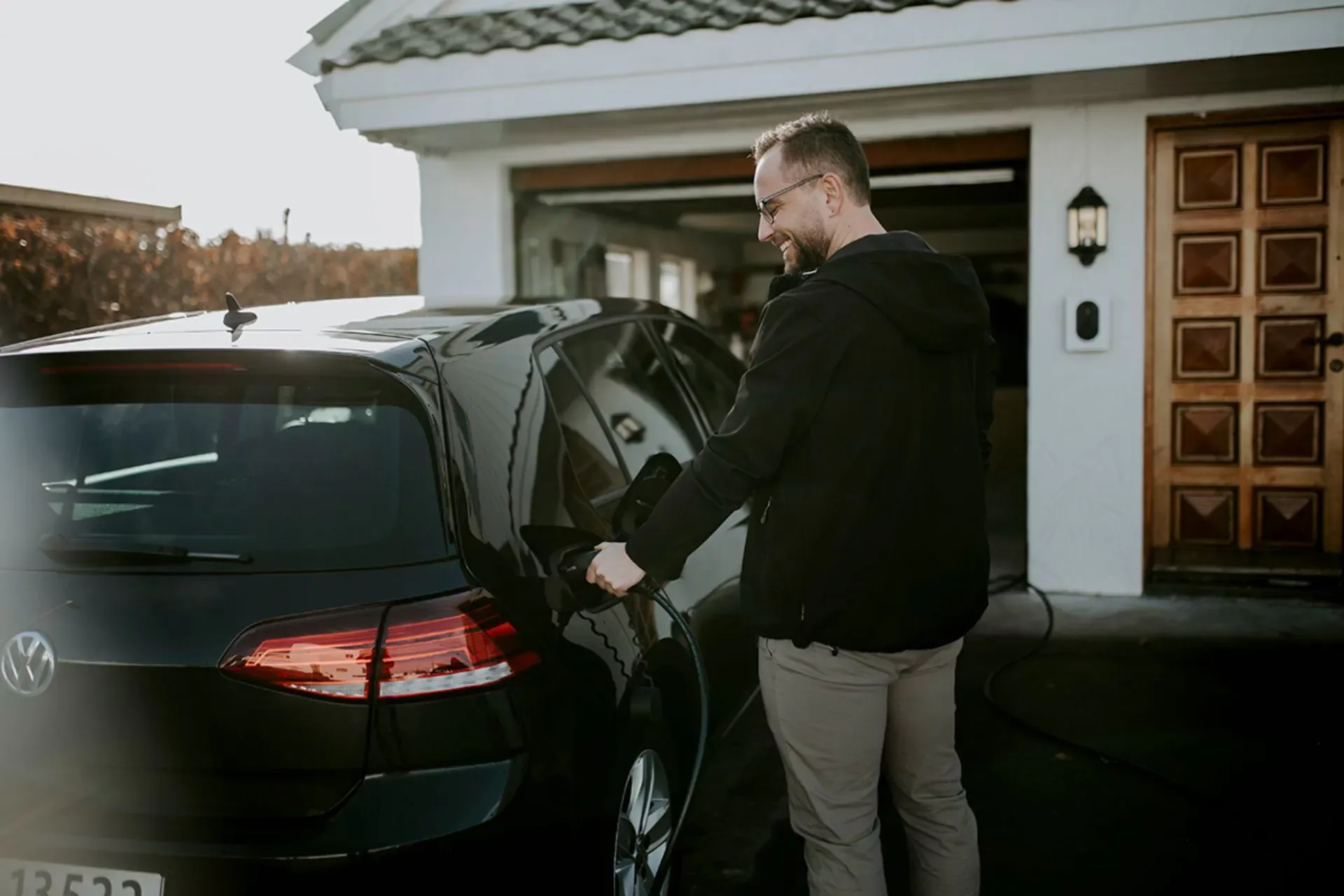 En mann smiler mens han lader en svart Volkswagen elbil med en hvit Zaptec Go montert på veggen ved inngangsdøren til et hvitt hus med åpen garasje.