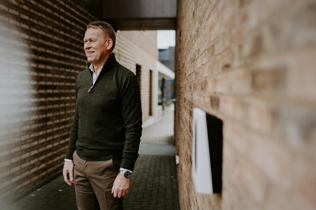 Kurt Østrem VD Zaptec står klädd i kläder i mörka naturnära toner intill en ljust brun tegelvägg, på väggen sitter en vit Zaptec Go laddare.