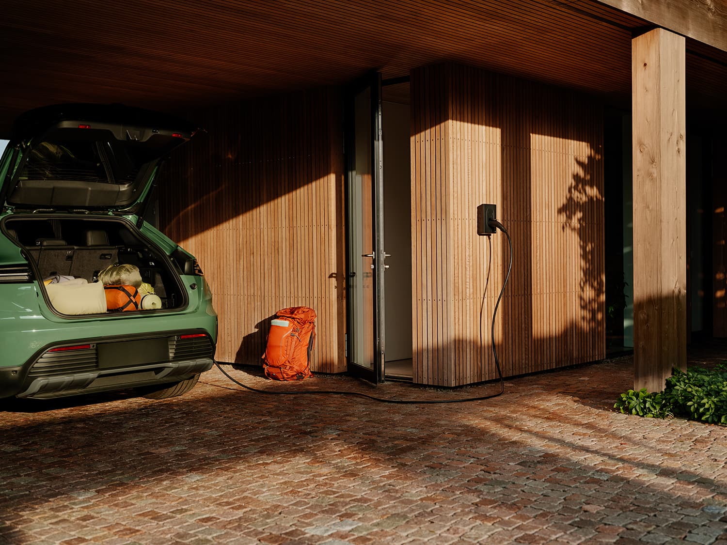 Voiture verte coffre ouvert en charge sur Zaptec Go borne mur bois, sac à dos orange à côté, sous carport moderne.
