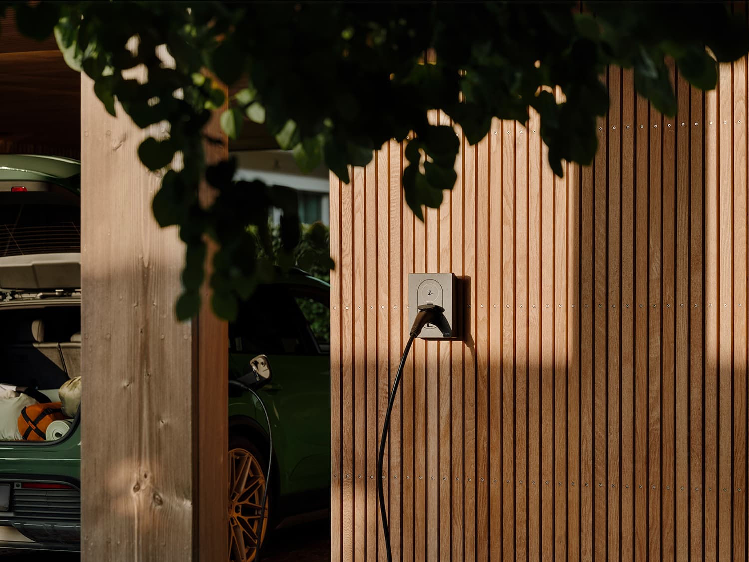 Zaptec Go EV charger on wooden wall, green car with open trunk and gold wheels partly visible, leafy branch above in foreground.
