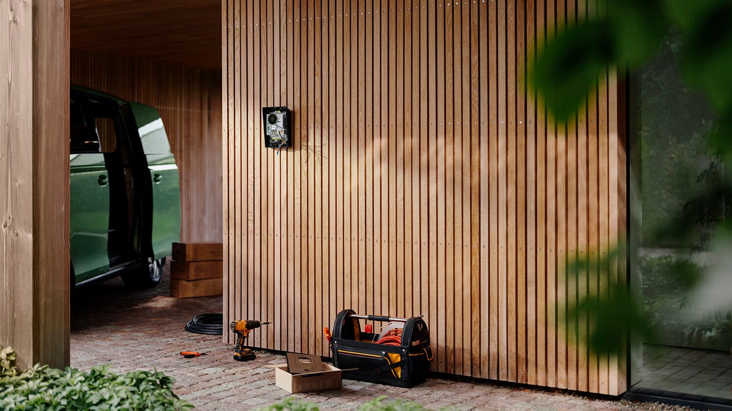 Zaptec Go 2 home charger installation in progress on wooden wall, green car with open door, tool bag and drill on cobblestone ground.