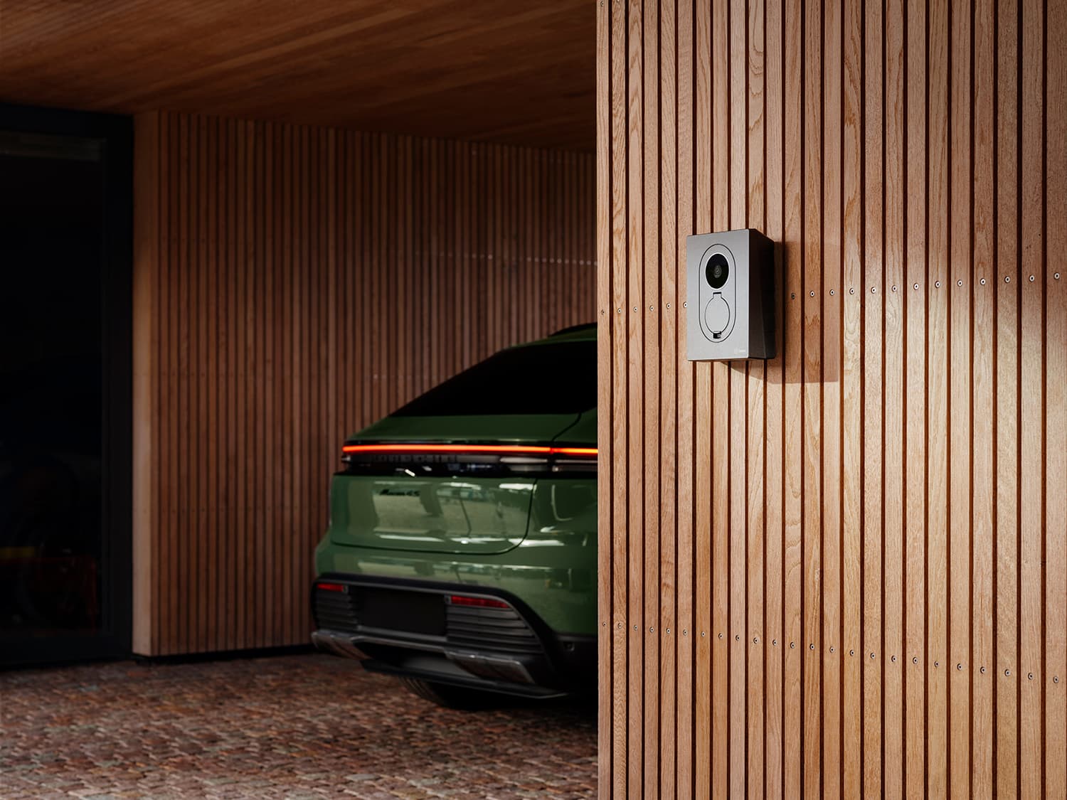 Bakenden av grønn bil i carport med Zaptec Go 2 hjemmelader på trevegg, sollys kaster skygger over området.