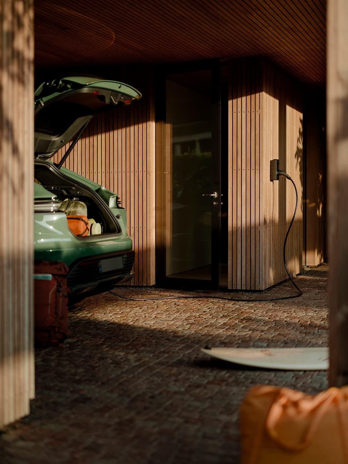 Green car with open trunk charging at Zaptec Go 2 home charger on wooden wall, orange backpack and surfboard in foreground.