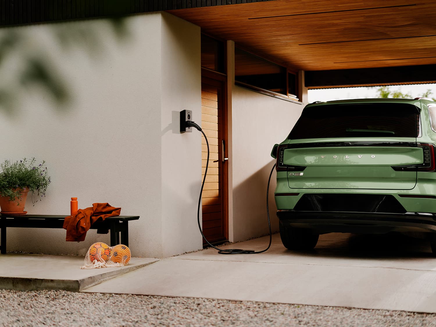 Bakenden av grønn Volvo lader med Zaptec Go 2 hjemmelader på hvit vegg, benk med oransje jakke og fotballer ved siden av.