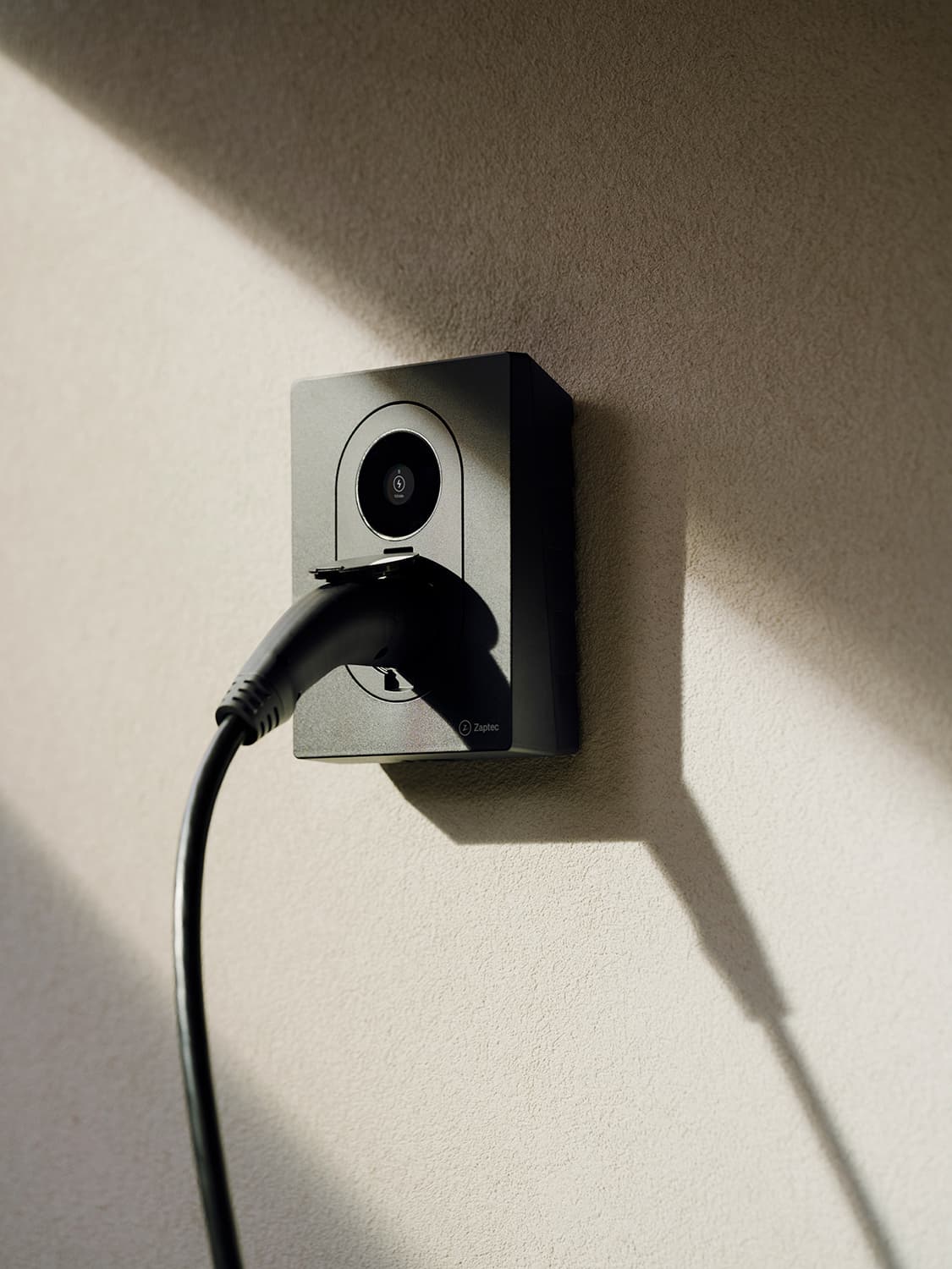 Close-up of Zaptec Go 2 home charger plugged in on white wall, strong shadow and sunlight cast across the surface.