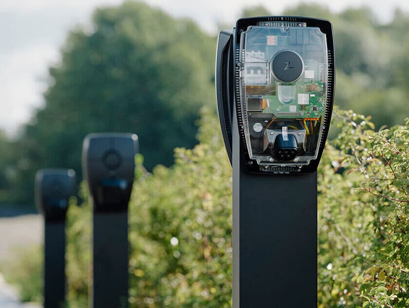 Nahaufnahme Zaptec Pro Ladestation auf Säule im Parkplatz, weitere Ladestationen auf Säulen im Hintergrund, Grünflächen.