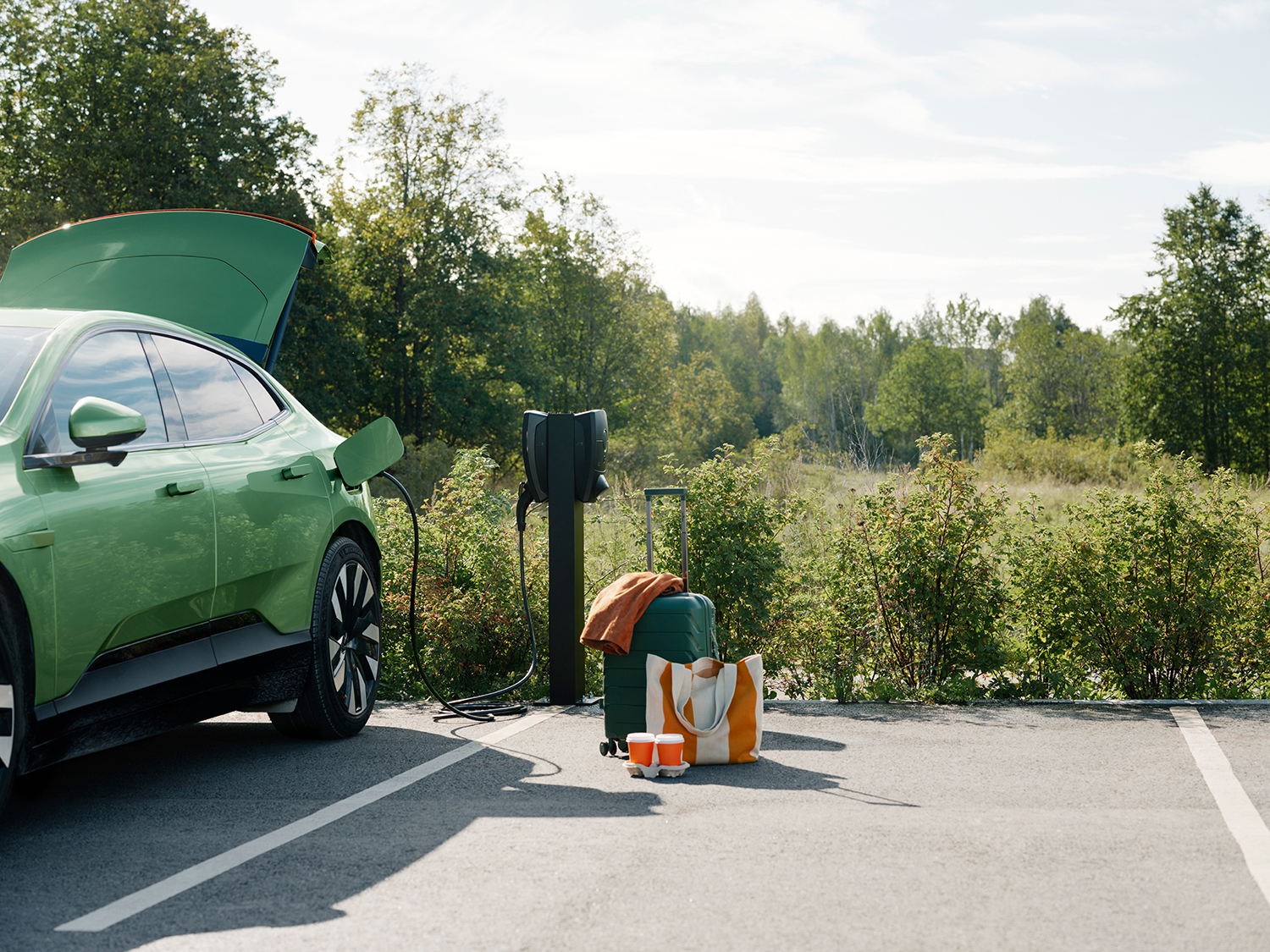 Groene auto laadt op bij Zaptec Pro lader op kolom op parkeerplaats, koffer, tas en bekers op de grond, open kofferbak.