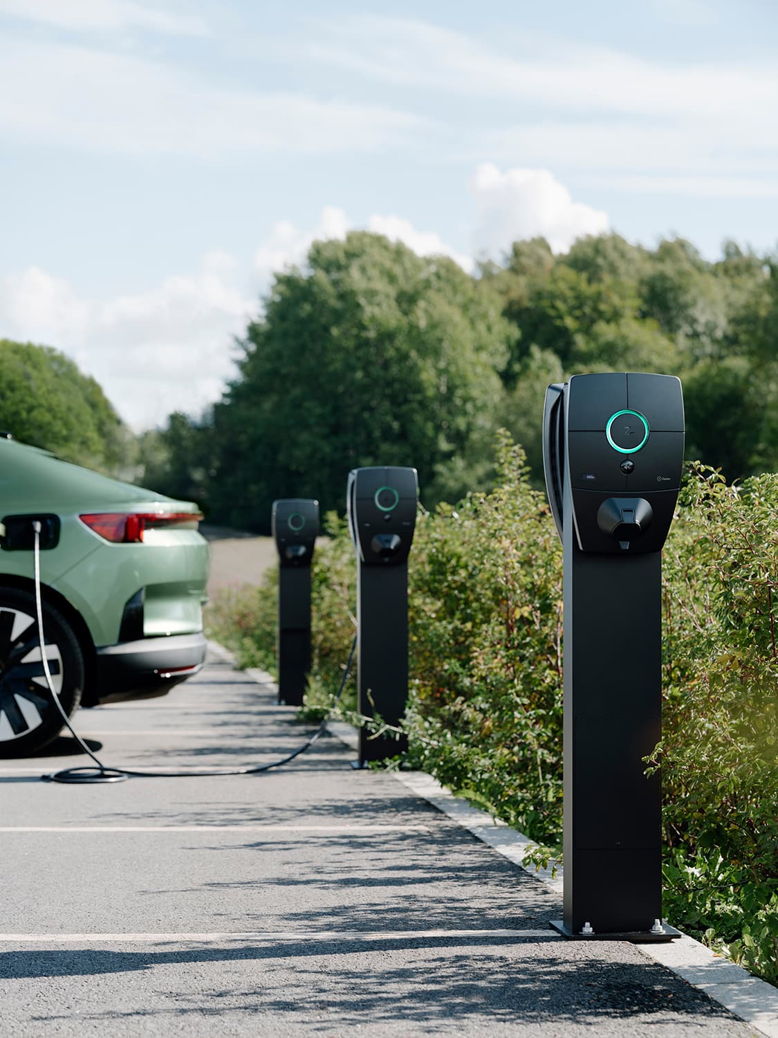 Three Zaptec Pro chargers mounted on columns in parking lot, green car charging at first charger, greenery in background.
