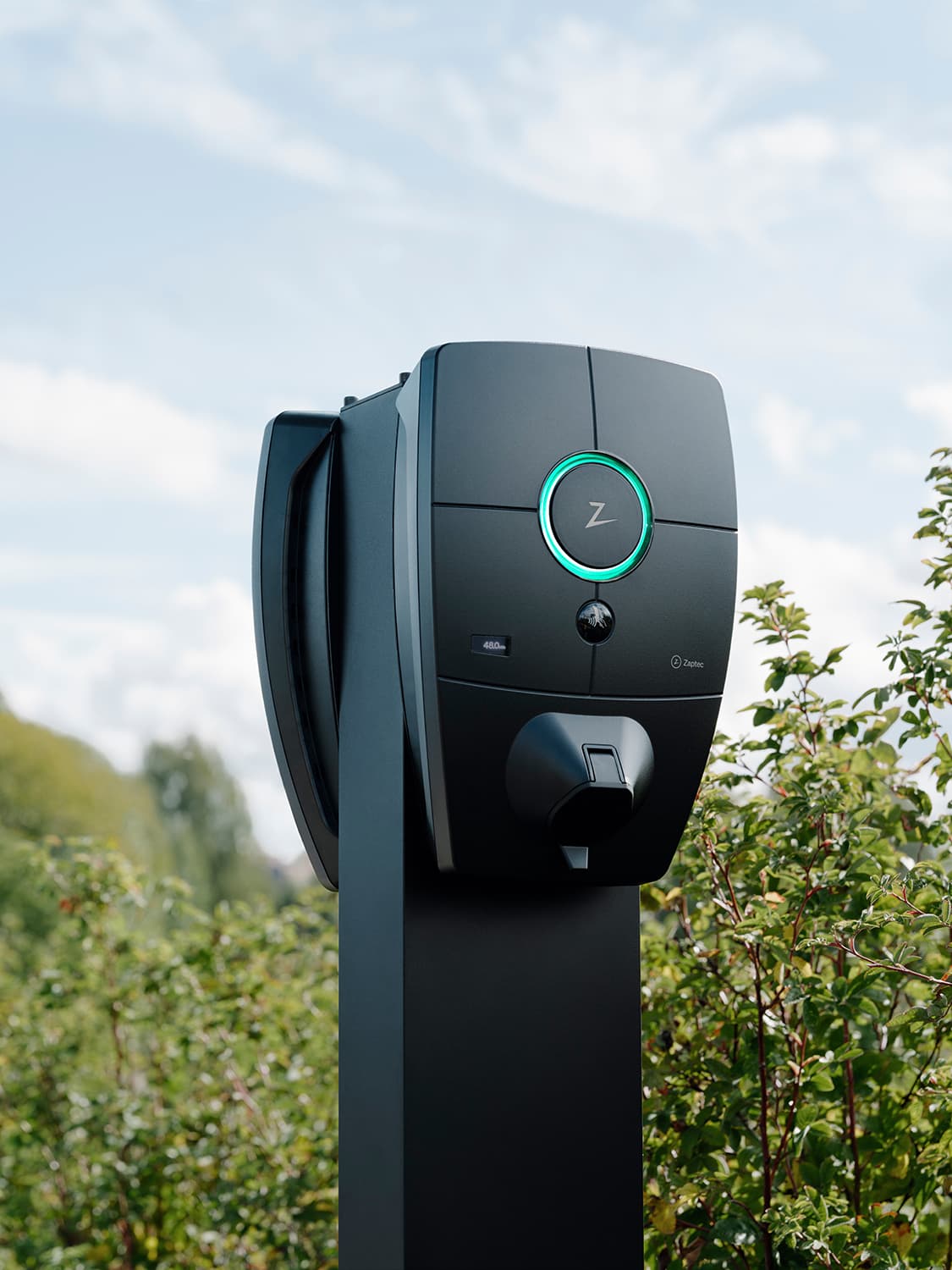 Close-up of Zaptec Pro charger mounted on column in parking lot, green light ring, greenery and blue sky in background.