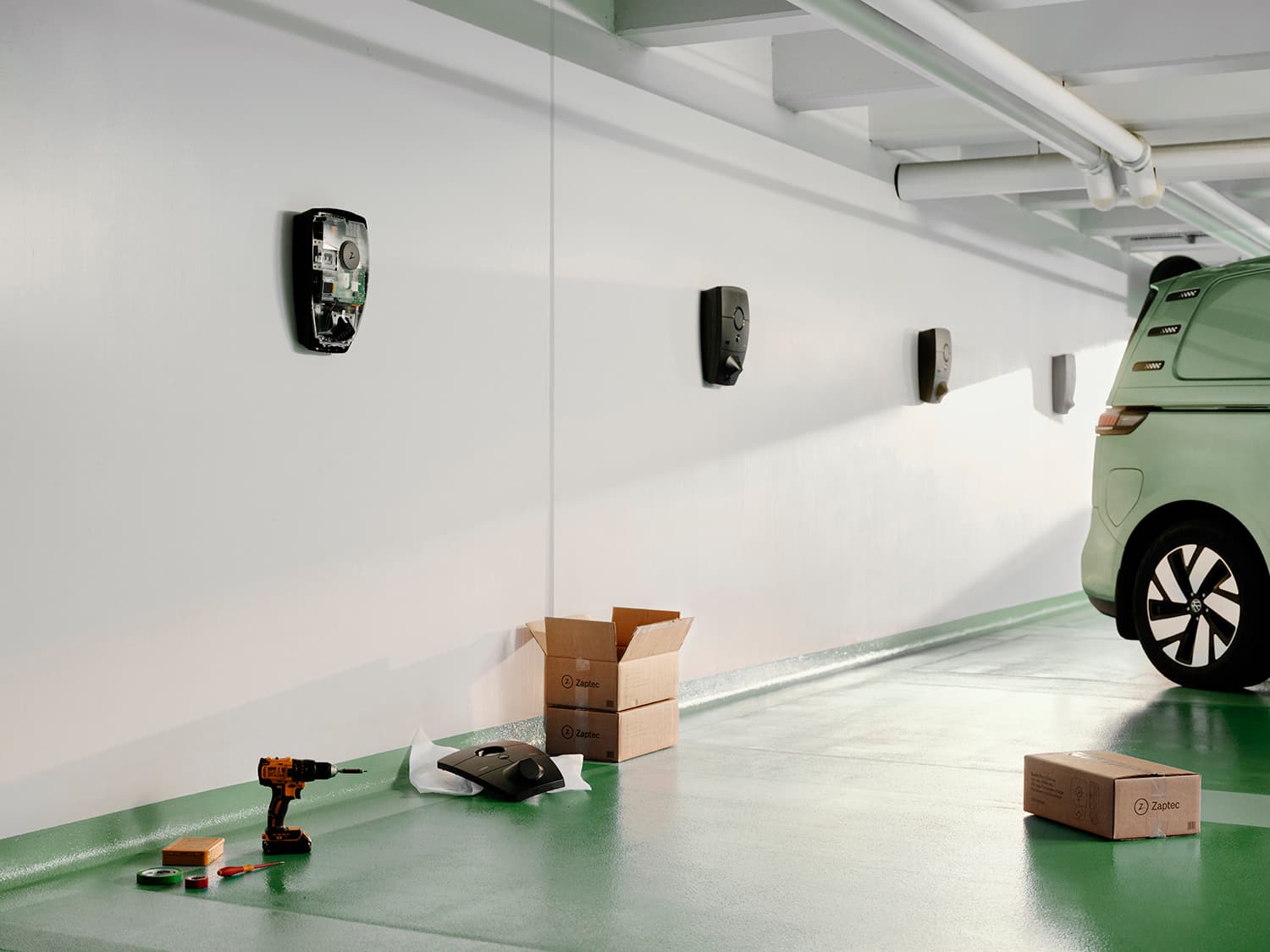 Meerdere Zaptec Pro laders op witte muur in parkeergarage, groene bestelwagen, boormachine, kartonnen dozen en afdekking op vloer.