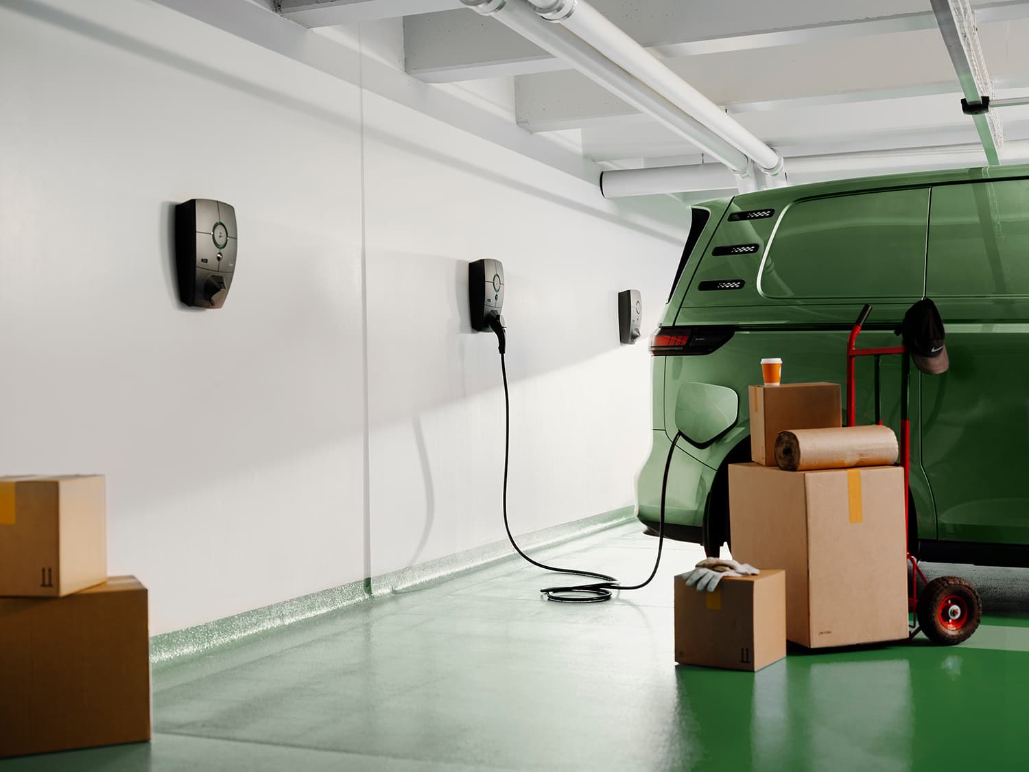 Groene bestelwagen laadt op met Zaptec Pro lader op witte muur in parkeergarage, gestapelde kartonnen dozen, handschoenen en koffiebeker op de voorgrond.