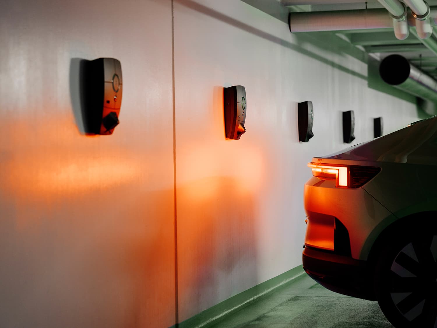 Plusieurs bornes Zaptec Pro sur mur blanc en parking, feu arrière rouge d’une voiture verte reflété sur le mur, lumière dramatique.