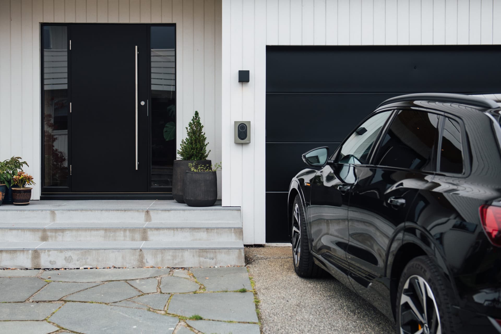 En svart elbil står parkert foran et hvitt hus med svart dør og garasjeport. En Rock Grey Zaptec Go-lader er montert på fasaden mellom døren og garasjen.