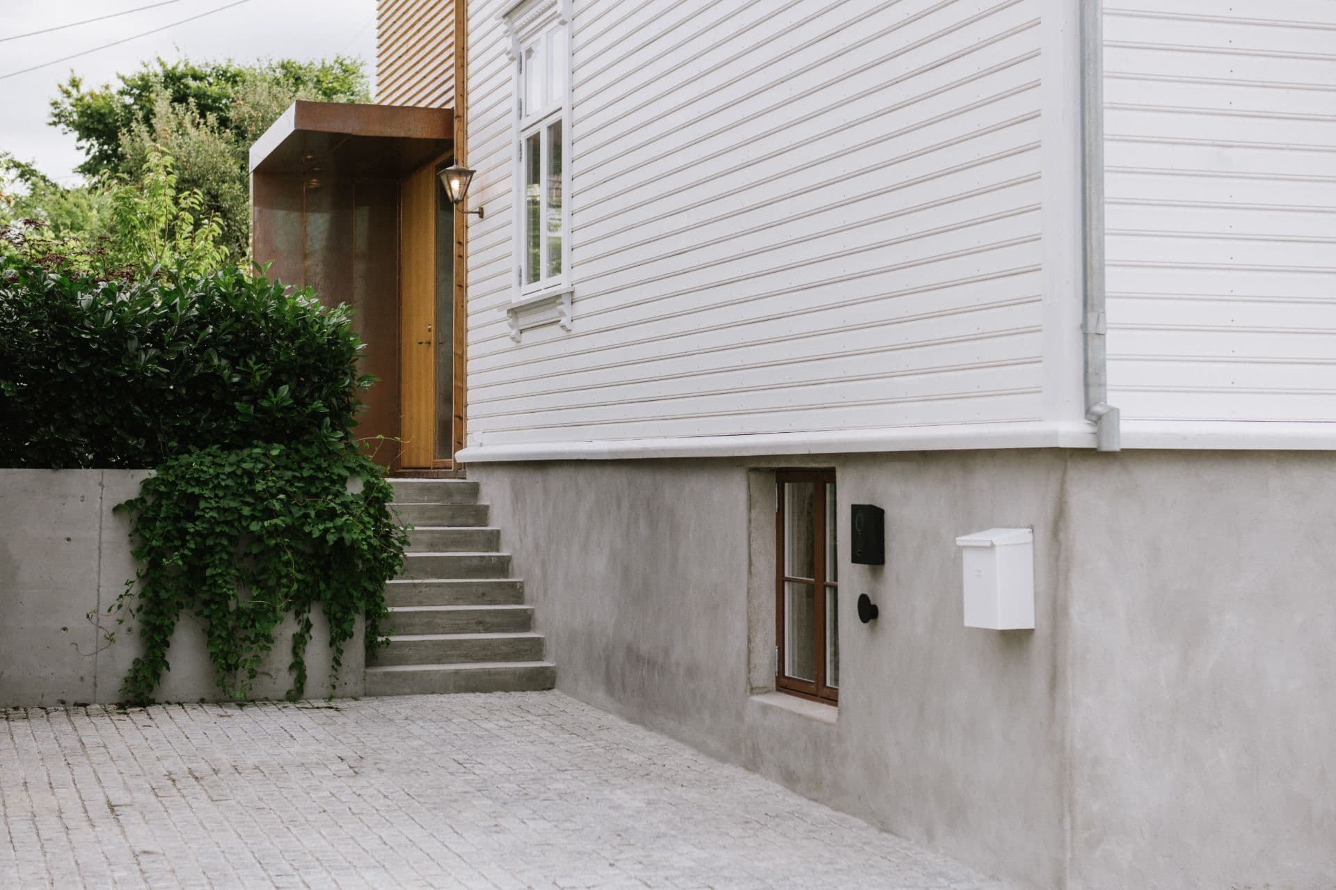 En svart Zaptec Go-lader er montert på betongfundamentet til et trehus med hvit kledning. En liten trapp fører opp til inngangsdøren, omgitt av grøntområder.