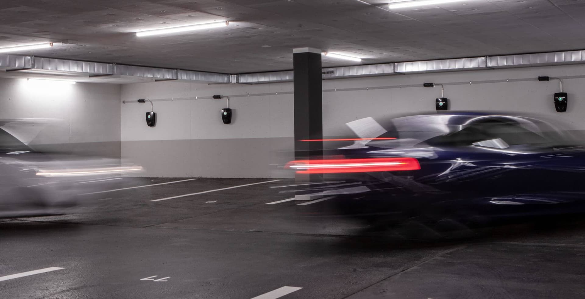 ett parkeringsgarage med flera elbilsladdare monterade på en vägg, två bilar ses swisha förbi.