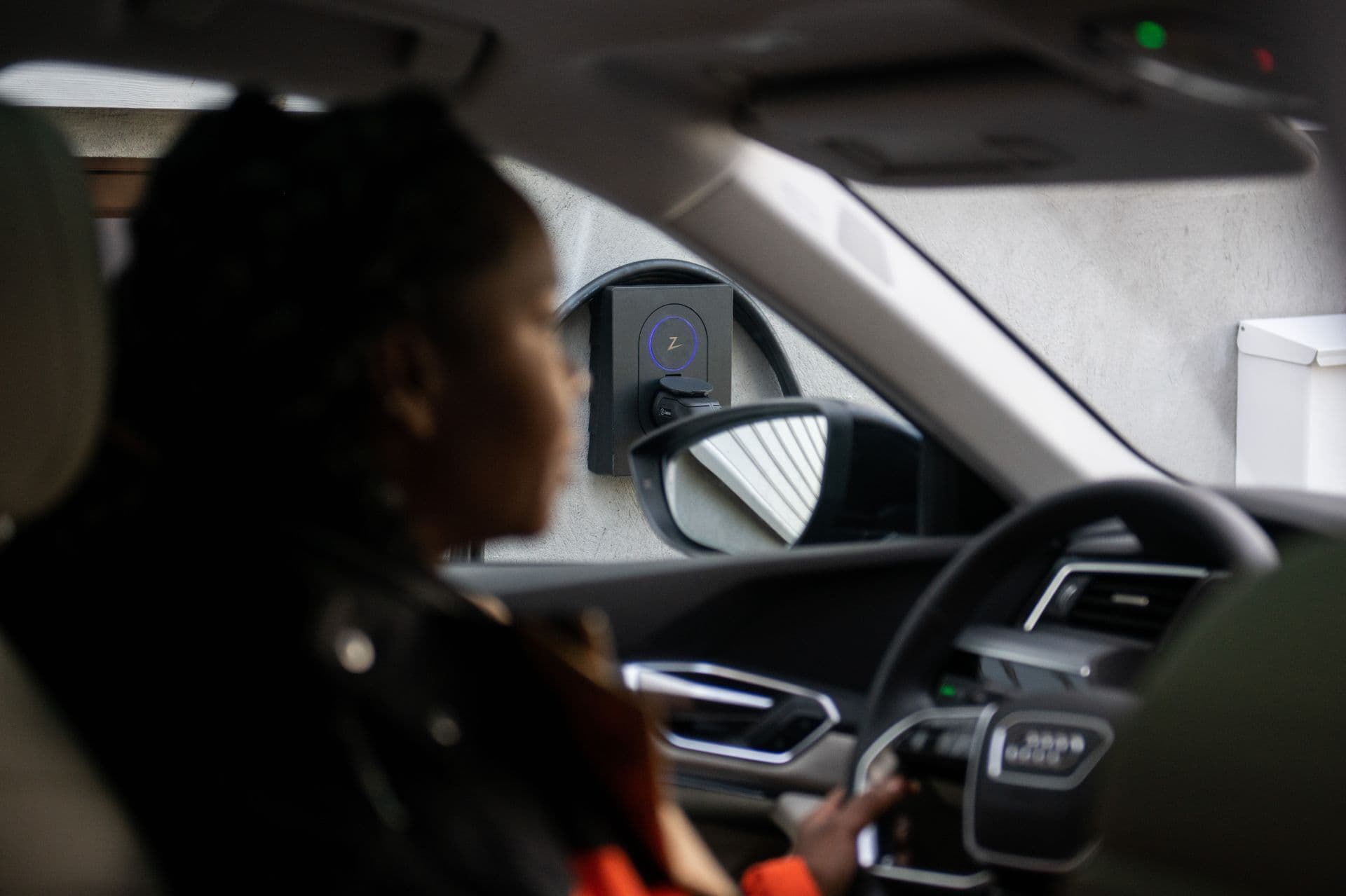En kvinne sitter i førersetet på en elbil og ser mot en Zaptec Go-lader montert på en lys vegg utenfor bilen. Laderen reflekteres også i sidespeilet.