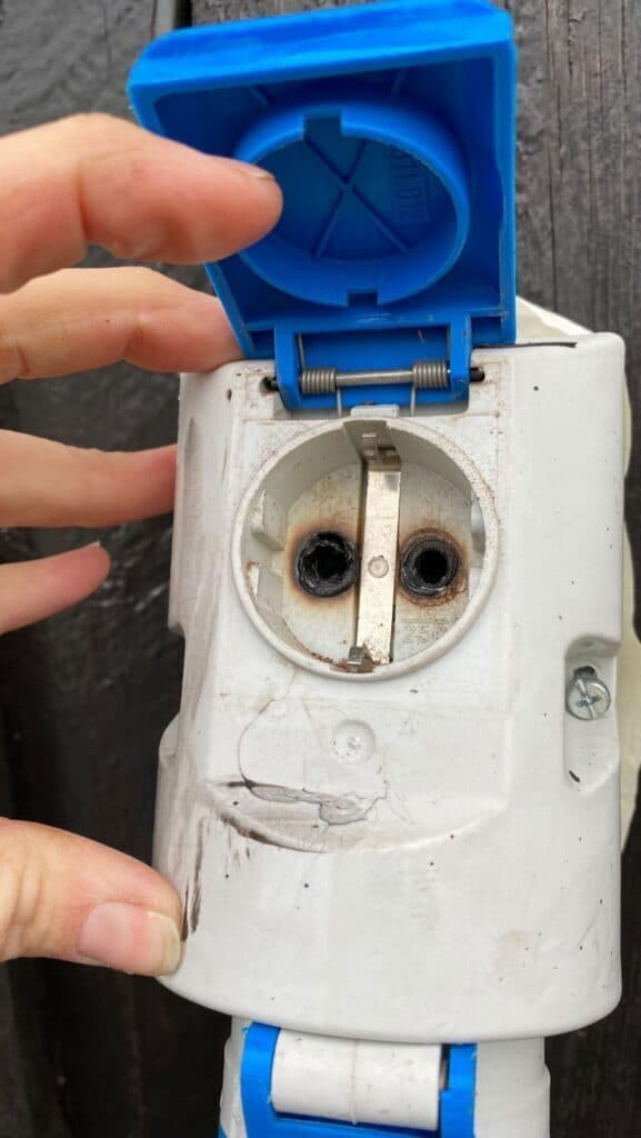 En nærbilde av en utendørs stikkontakt med blått lokk som holdes åpent av en hånd. Kontakten er misfarget og svidd rundt hullene, og det er synlige brennmerker og sprekker i plasten.