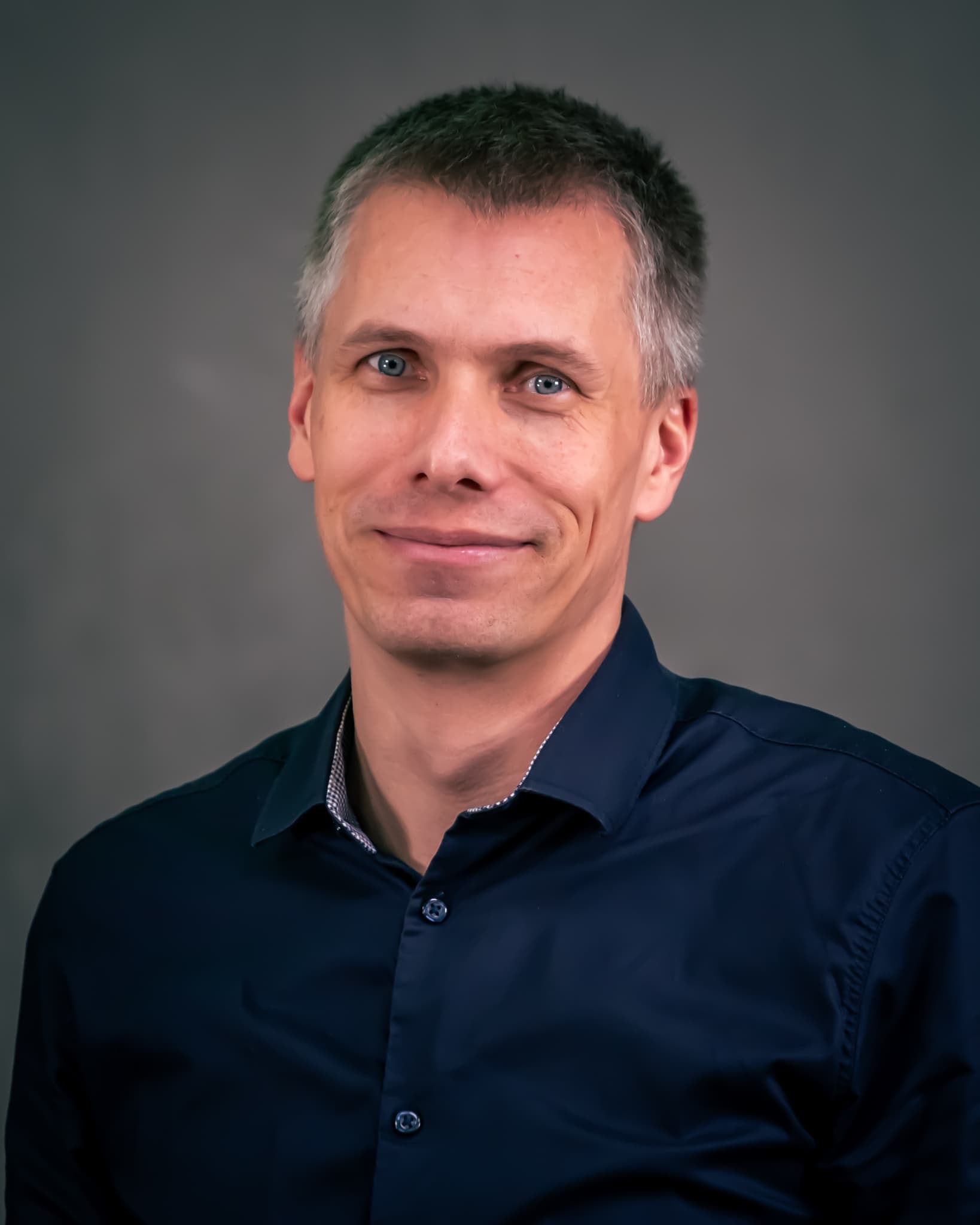 Portrett av en mann med kort brunt hår og blå skjorte, som smiler svakt foran en nøytral, mørk bakgrunn.