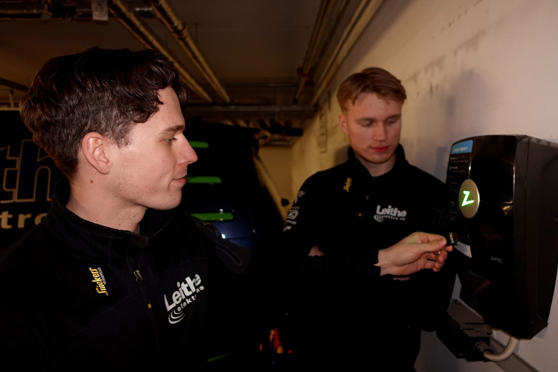 To elektrikere fra Leithe elektro står i en garasje og tester en Zaptec Pro-lader som er montert på en vegg. Den ene holder opp et RFID-kort mot laderen.