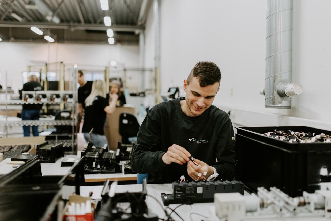 En person jobber med montering av elektroniske komponenter ved en arbeidsstasjon i et lyst fabrikklokale og smiler konsentrert.