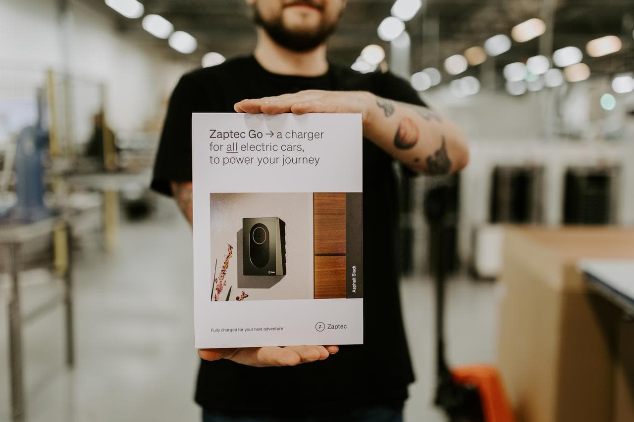 En person i svart genser holder opp en eske med en Zaptec Go-lader i et fabrikkmiljø. Eskens framside viser et bilde av laderen montert på en trefasade.