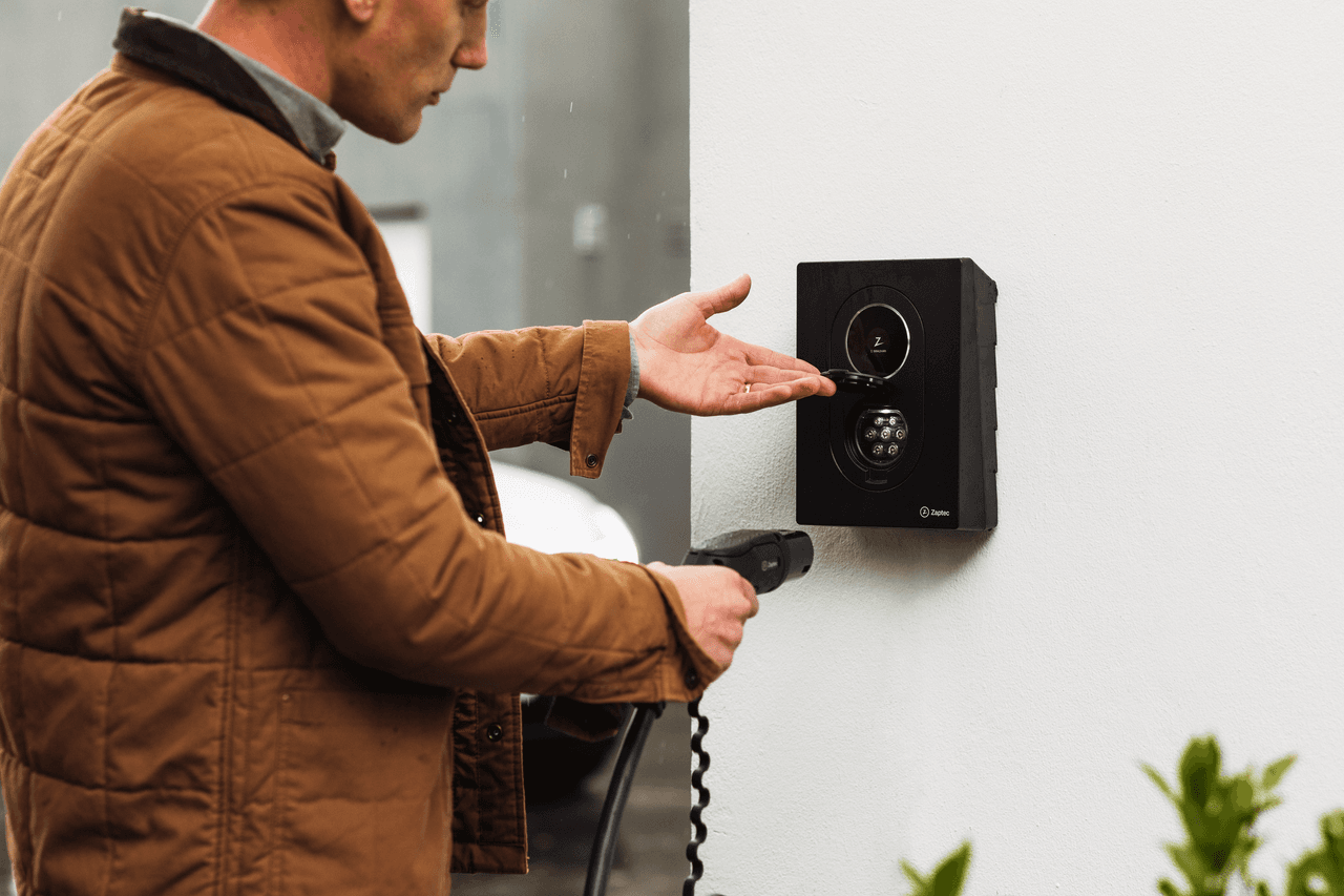 En man i en brun jacka håller en laddkabel i ena handen och lyfter locket på en svart Zaptec Go 2 laddare som är monterad på en vit vägg.