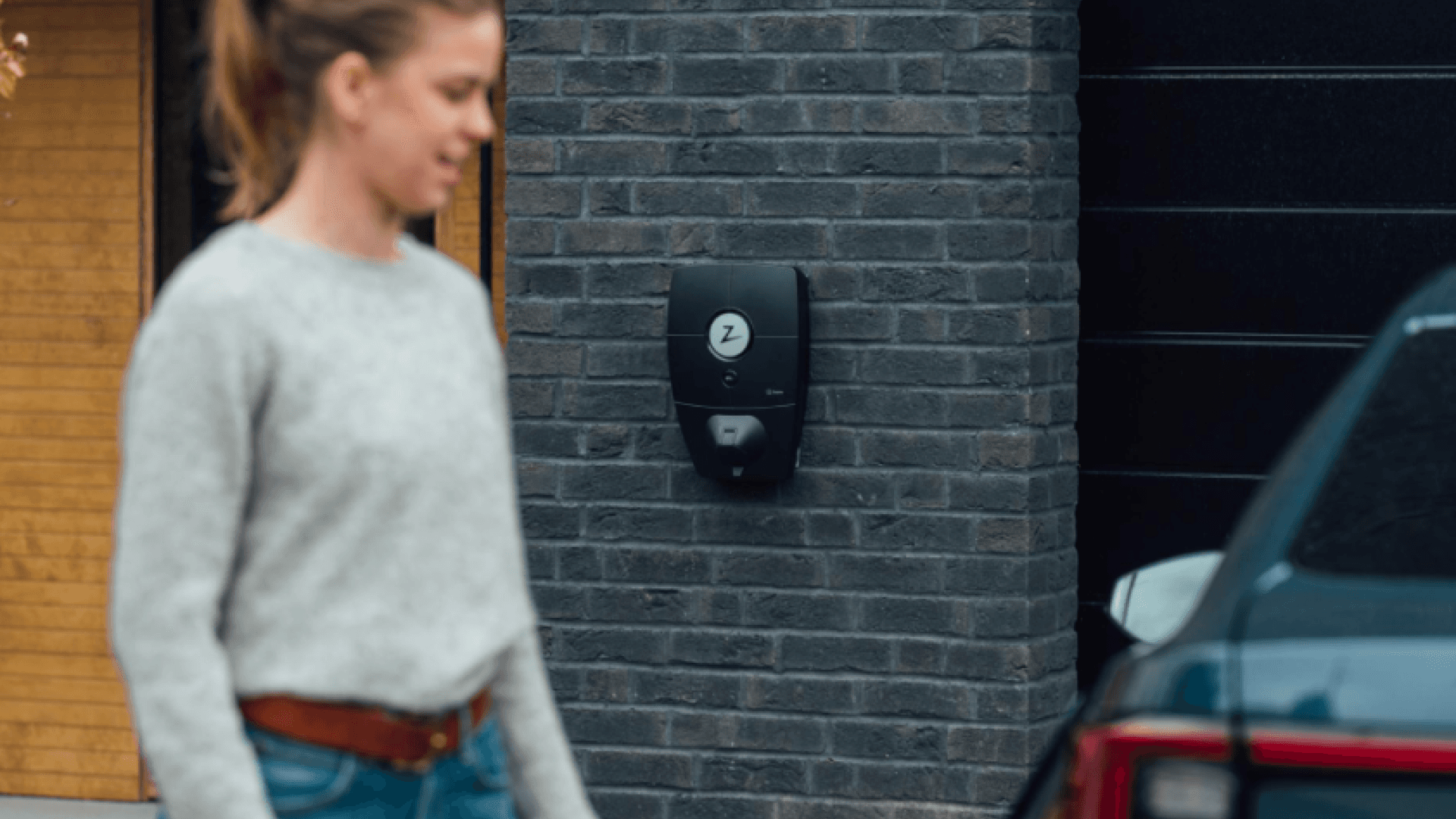 En kvinna i grå tröja går mot en mörk elbil, på en mörkt grå tegelvägg mellan henne och bilen sitter en Zaptec Pro laddbox