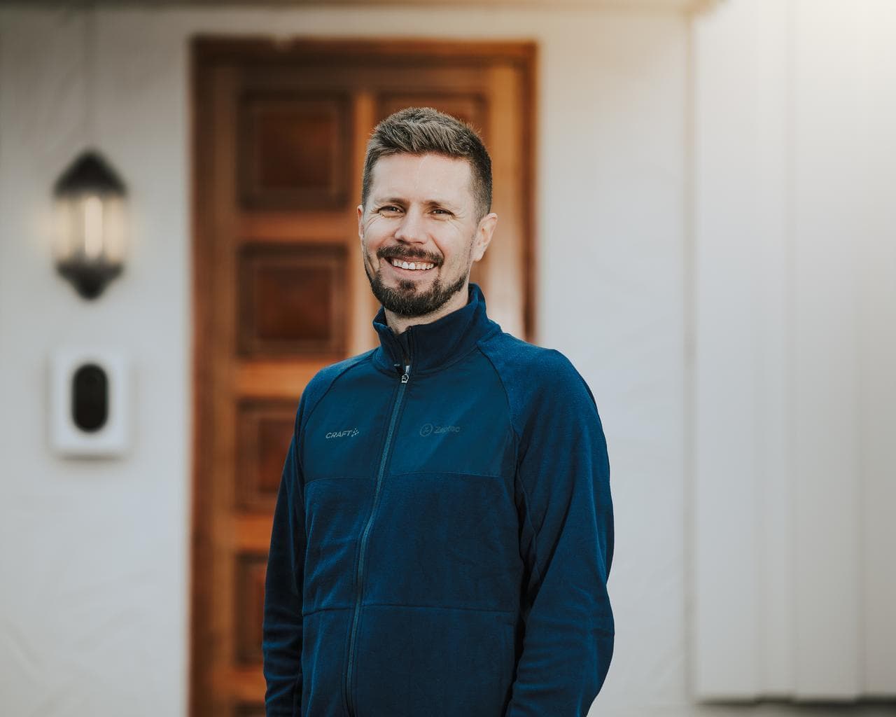 En smilende mann i blå jakke står foran en ytterdør i tre, med en hvit Zaptec Go-lader montert på veggen ved siden av en utelampe.