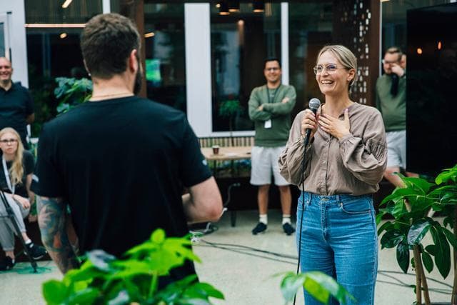 en kvinna med beige blus och jeans håller en mikrofon i ena handen och den andra handen håller hon högt upp på bröstet runt henne står och sitter personer och lyssnar, gröna växter syns i förgrunden