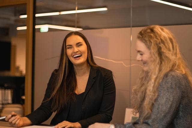 en skrattande kvinna med mörkt hår och mörk kavaj sitter vid ett skrivbord tillsammans med en kvinna i lockigt ljust hår och grå kavaj