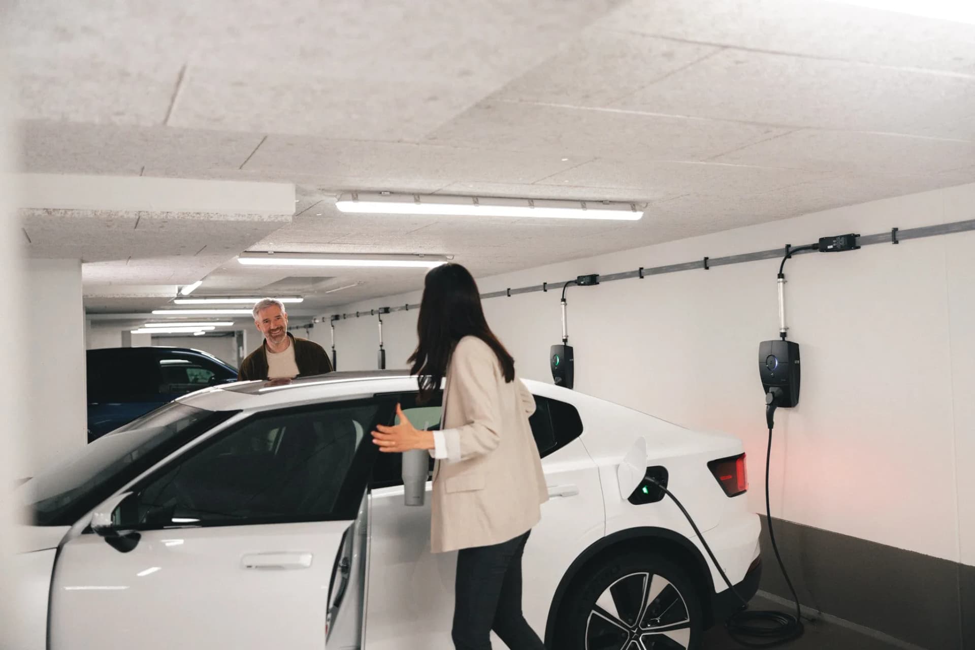En kvinnar laddar en vit elbil medan en man närmar sig och ler. Flera zaptec pro laddboxar syns monterade längs väggen i bakgrunden.